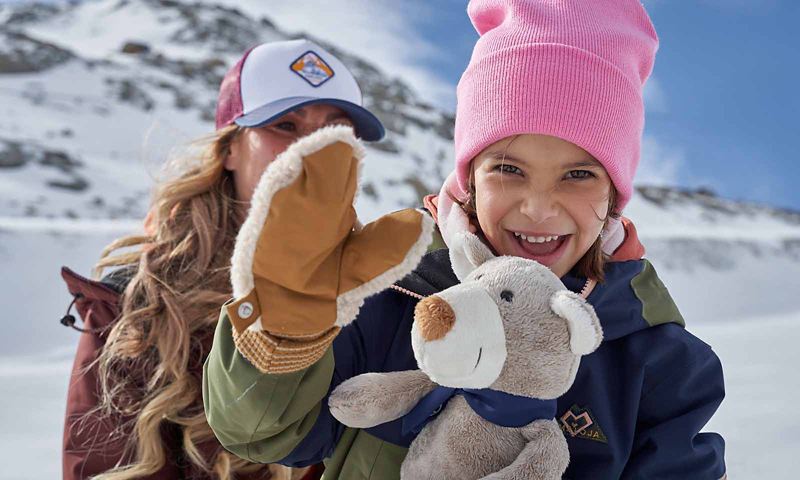 Una bambina con guanti e cappello di lana, in braccio alla madre, saluta sorridente in camera. Sullo sfondo montagne innevate.