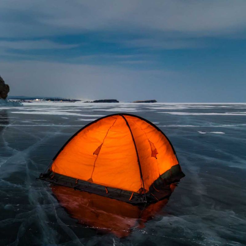 Una tenda da campeggio illuminata dall'interno, montata sulla superficie di un lago ghiacciato; sullo sfondo un promontorio roccioso affiora dal ghiaccio.