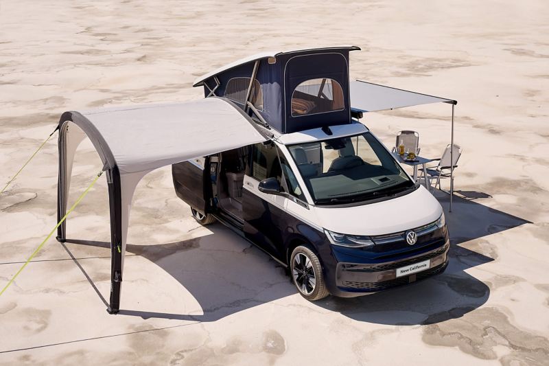 Photo of a VW California parked on a beach with its roof unit and awning deployed. 