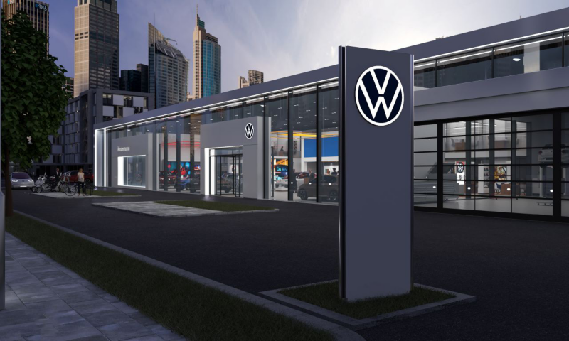 Une concession automobile Volkswagen moderne avec de grandes baies vitrées expose des véhicules à l'intérieur. Le logo Volkswagen est clairement visible sur un panneau extérieur.