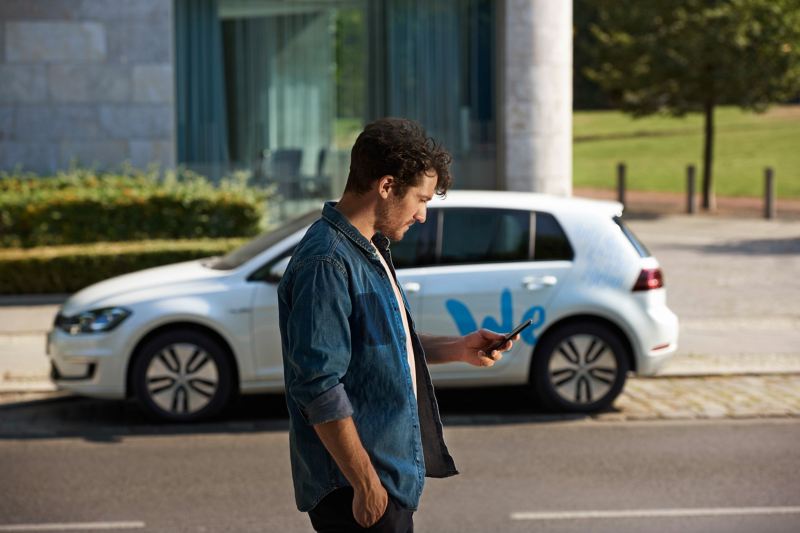 A man looking down at his phone stands in the foreground in front of an e-golf