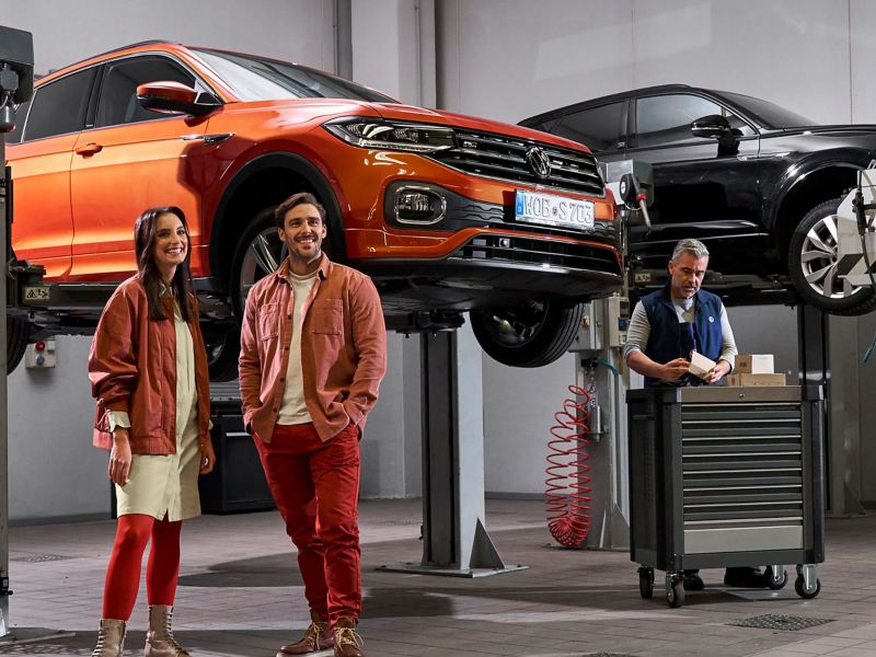 A couple standing in a VW repairs and service station with 2 cars on a ramp in the background