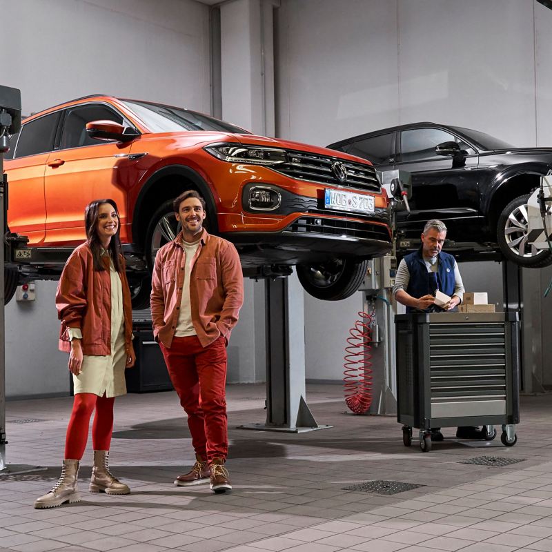 A couple standing in a VW repairs and service station with 2 cars on a ramp in the background