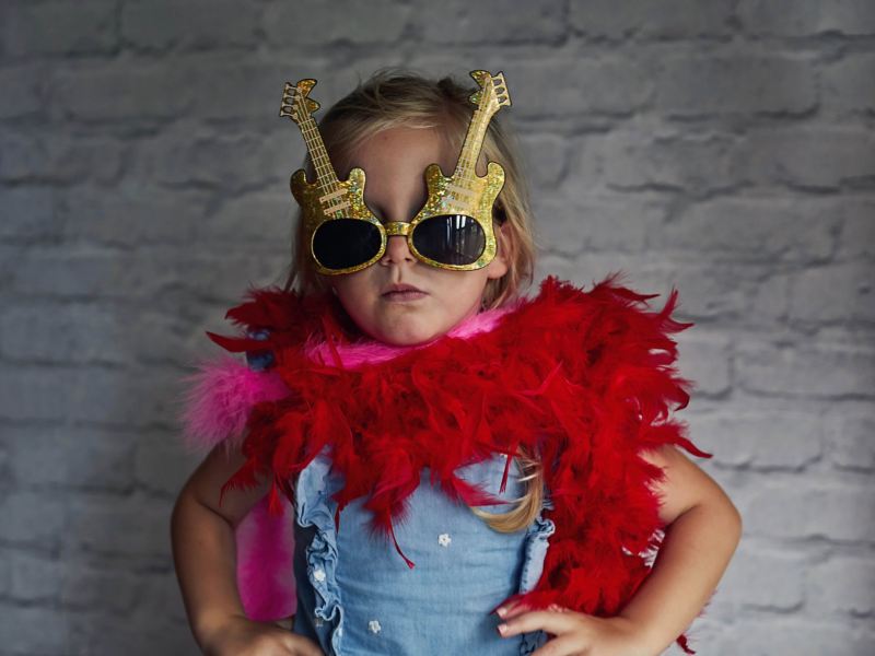 Confident little girl standing in front of a wall with sunglasses in the shape of a guitar 