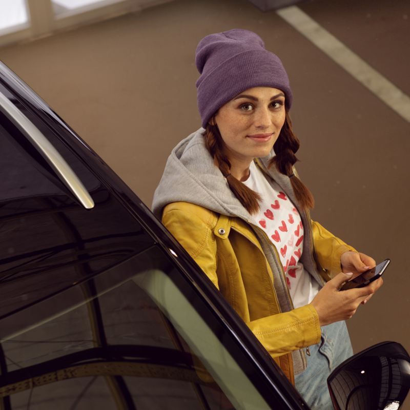 woman wearing a purple hat leaning against a car looking at the camera whilst holding a mobile phone