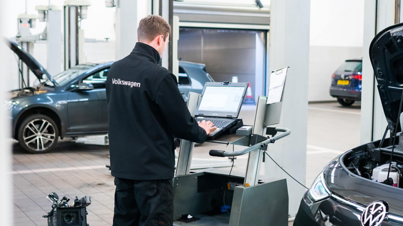 A Volkswagen mechanic in a Volkswagen jacket works on a laptop at a service center. Cars with open bonnets are in the background.