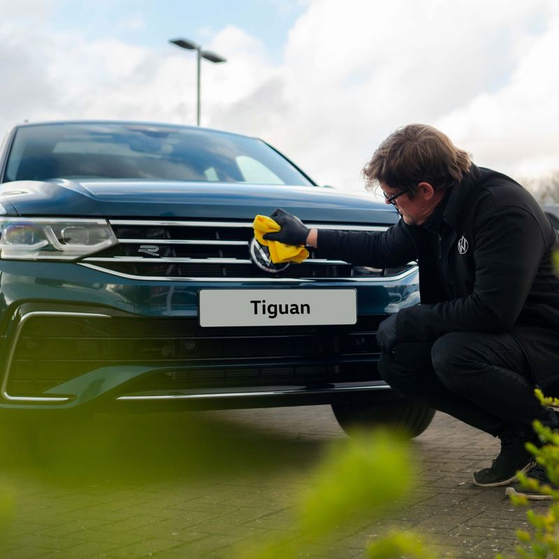 A service employee polishing the front badge on a blue VW Tiguan