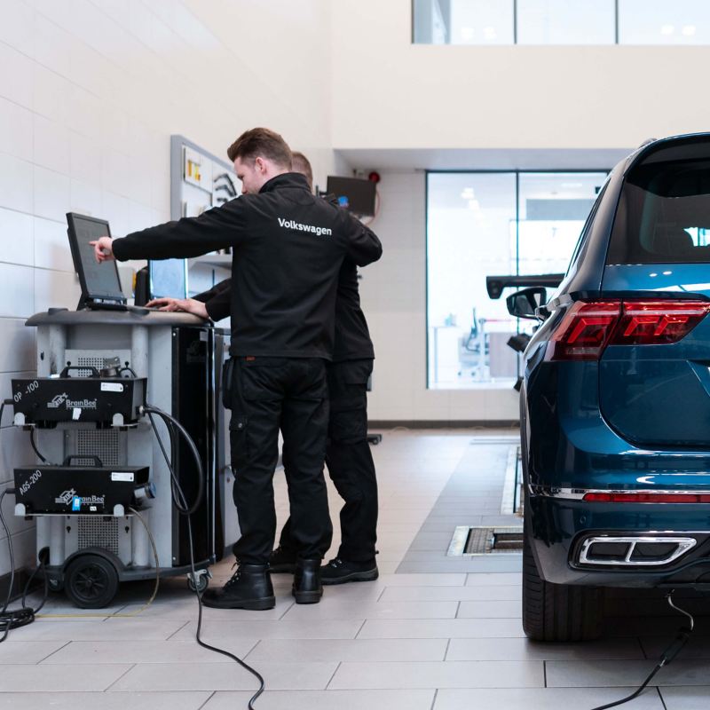 Two VW technicians stand at a computer with a blue VW Tiguan beside them