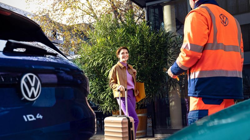 A person wheeling a suitcase being greeted by a VW roadside assistance technician