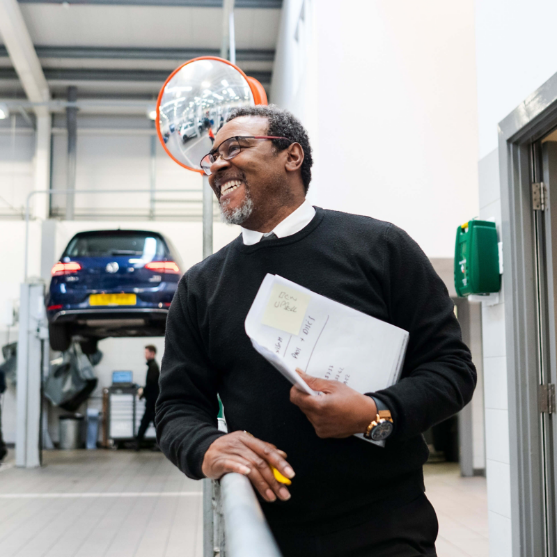 A service manager smiling holding paperwork with blue VW on ramp in background