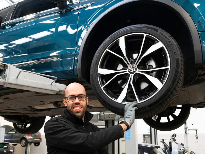A VW mechanic standing next to a raised car with his hand on the tyre
