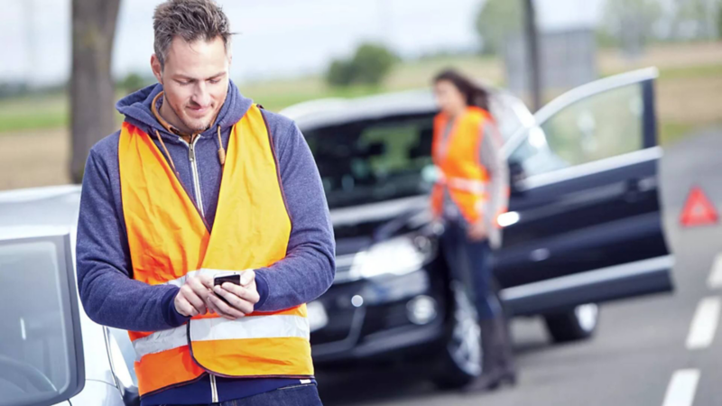 Conductor de Volkswagen con ropa de alerta vial color naranja pide ayuda con su celular. 