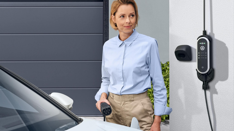 A woman in a light blue shirt holds an electric car charging plug, standing beside a Volkswagen Passat car. A Volkswagen charger is mounted on the wall, conveying modernity and eco-friendliness.