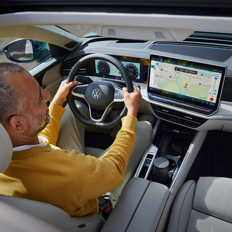 Man in a yellow sweater driving a Volkswagen Passat, focusing on the digital navigation screen on the dashboard. The interior is modern and sleek.
