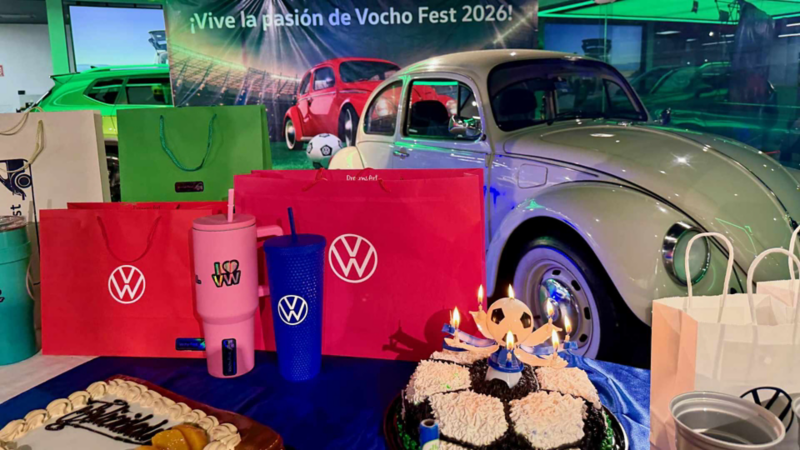 Pastel y vasos conmemorativos frente a Volkswagen Sedan Vocho de color blanco en evento.
