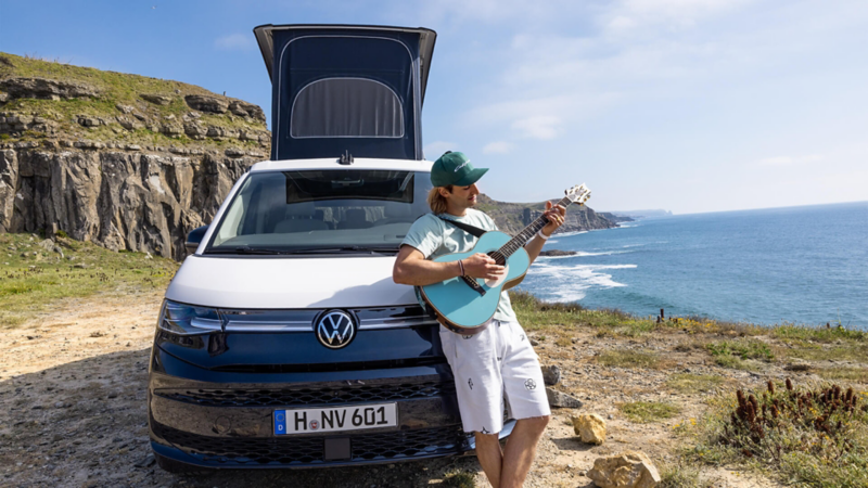 ein Mann lehnt gegen einen VW California und spielt Gitarre.