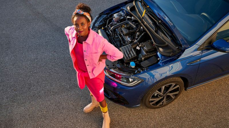 Une femme attends devant le capot ouvert de sa Volkswagen, dévoilant le moteur.