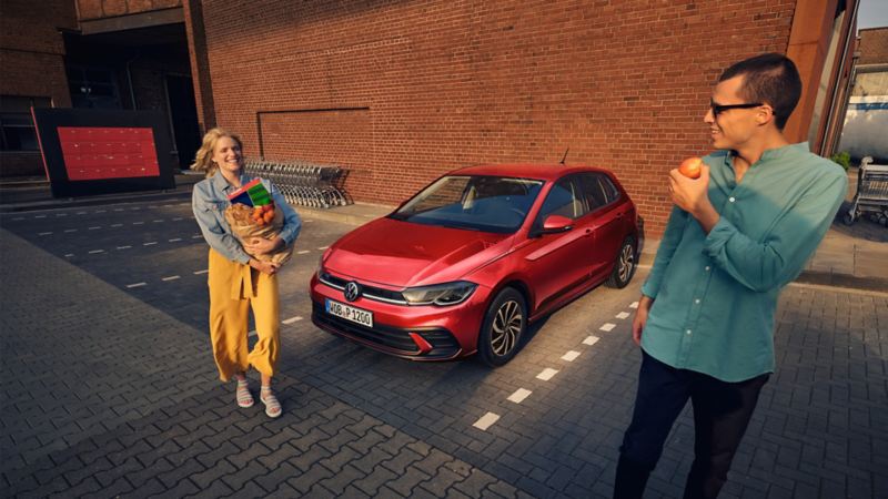 En rød Polo Life holder på en parkeringsplads, fronten set skråt forfra. En kvinde med en indkøbspose på armen går smilende forbi.