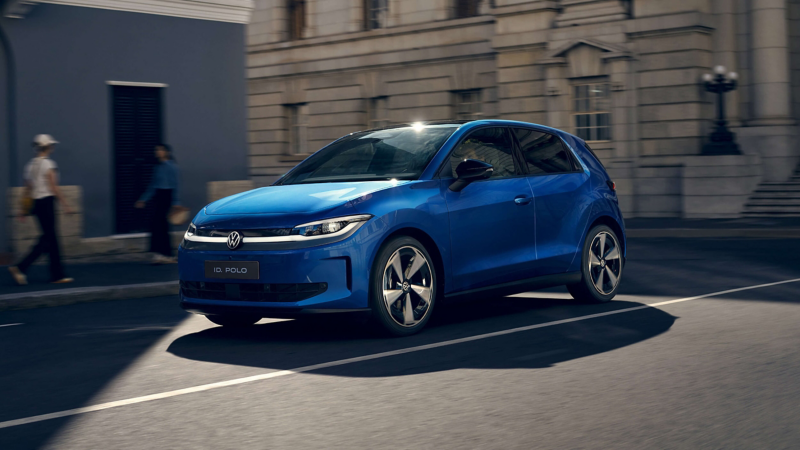 A sleek blue hatchback car is parked on a sunlit street, casting a shadow. Two pedestrians walk by a historic building, adding an urban feel.