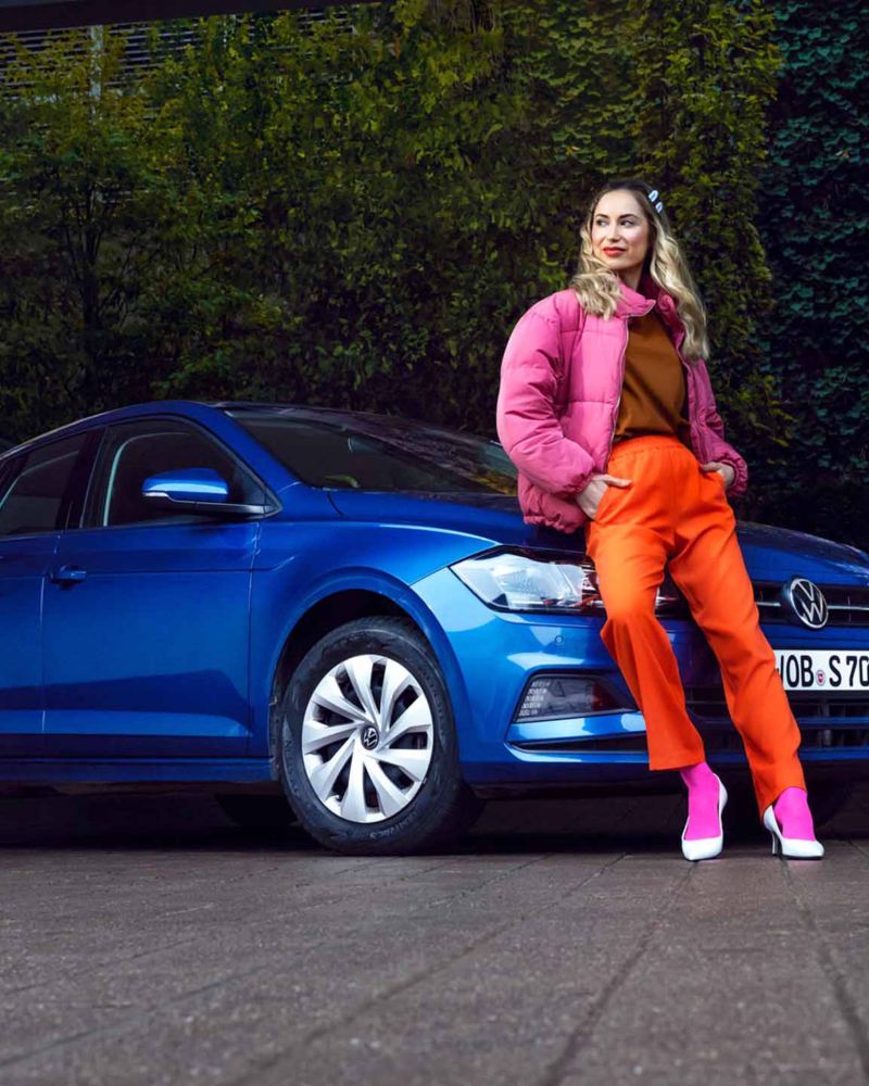 Una ragazza appoggiata al frontale di una Volkswagen Polo blu.
