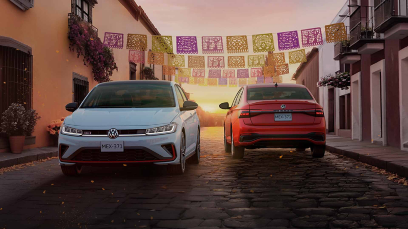 Dos sedanes Jetta, uno blanco de frente y otro rojo de espaldas, en una calle con papel picado.