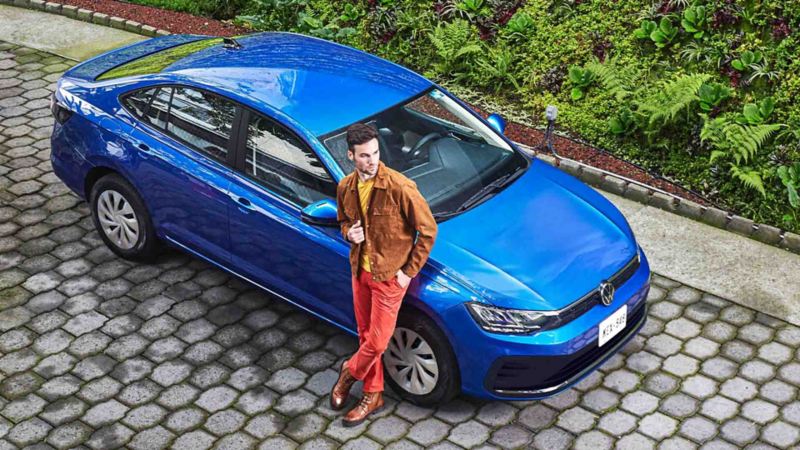Auto Virtus 2023 de Volkswagen en color azul, con joven conductor recargado cerca de puerta de copiloto. 