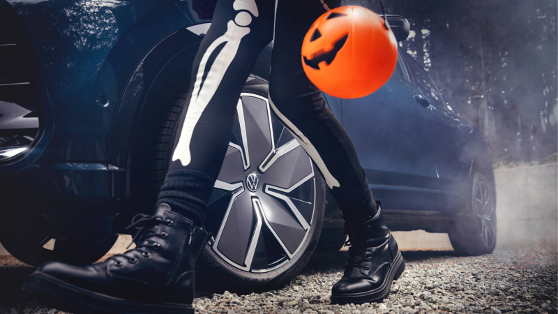 A person in black boots and skeleton-print leggings stands beside a dark car’s alloy wheel, holding a glowing orange jack-o’-lantern balloon in a Halloween scene.