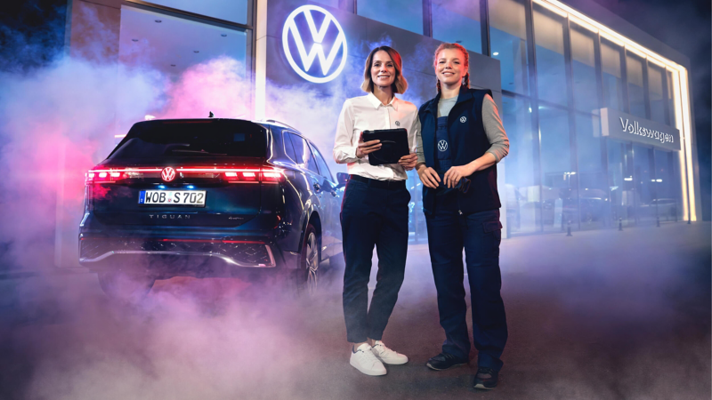 Two women stand beside a Volkswagen SUV amid pink and blue smoke outside a lit showroom, one holding a tablet.