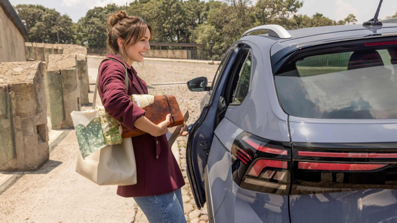 Mujer con varios bolsos junto a un SUV gris con la puerta trasera abierta, en un entorno arbolado.
