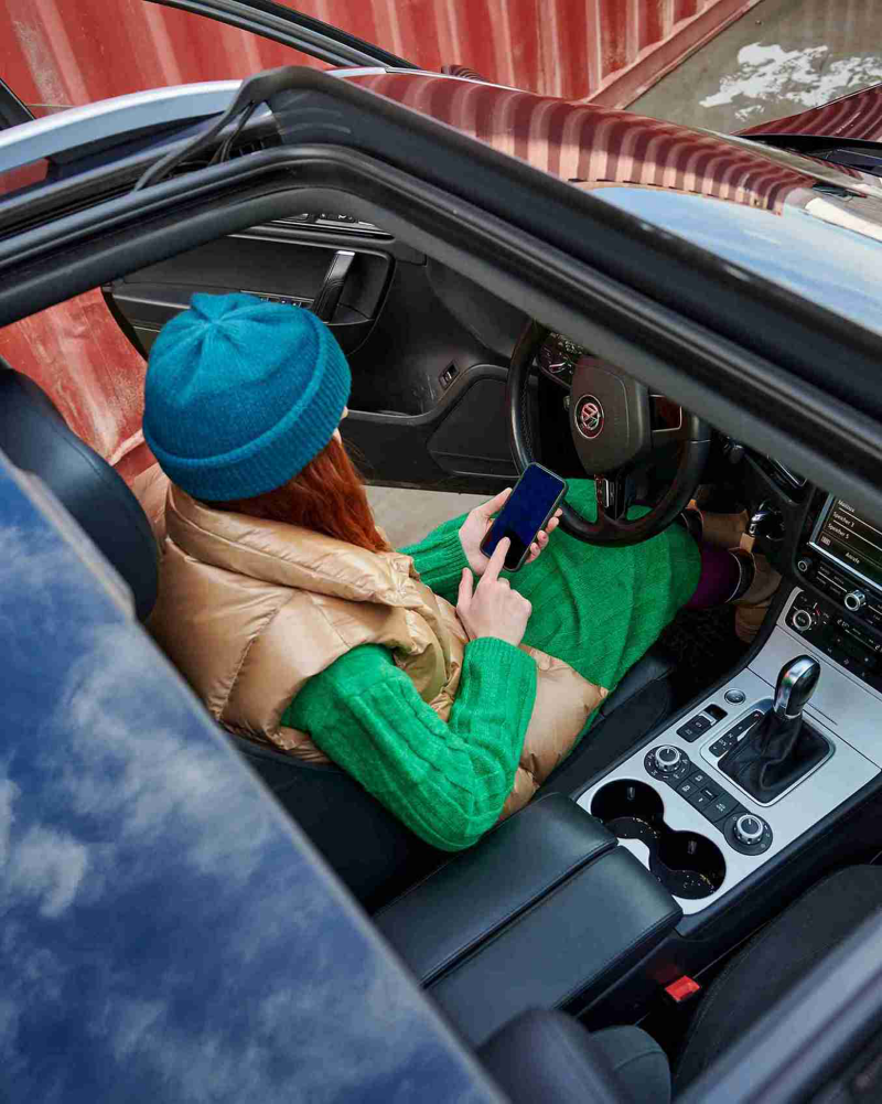 Mujer pelirroja en un coche usando un teléfono inteligente, con un gorro de punto azul y un suéter verde.
