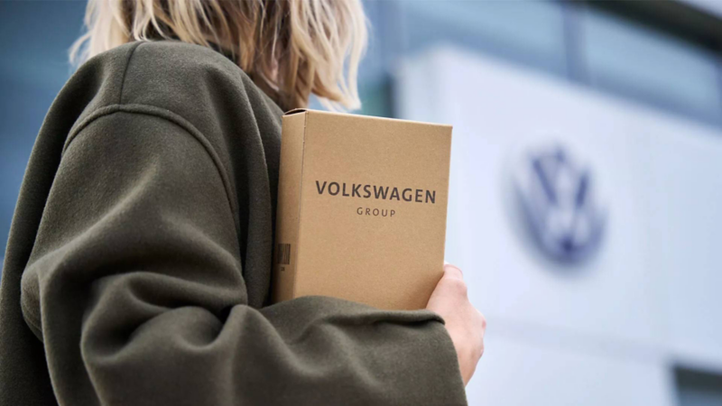 Mujer toma caja de refacciones Volkswagen mientras mira a una concesionaria Volkswagen. 