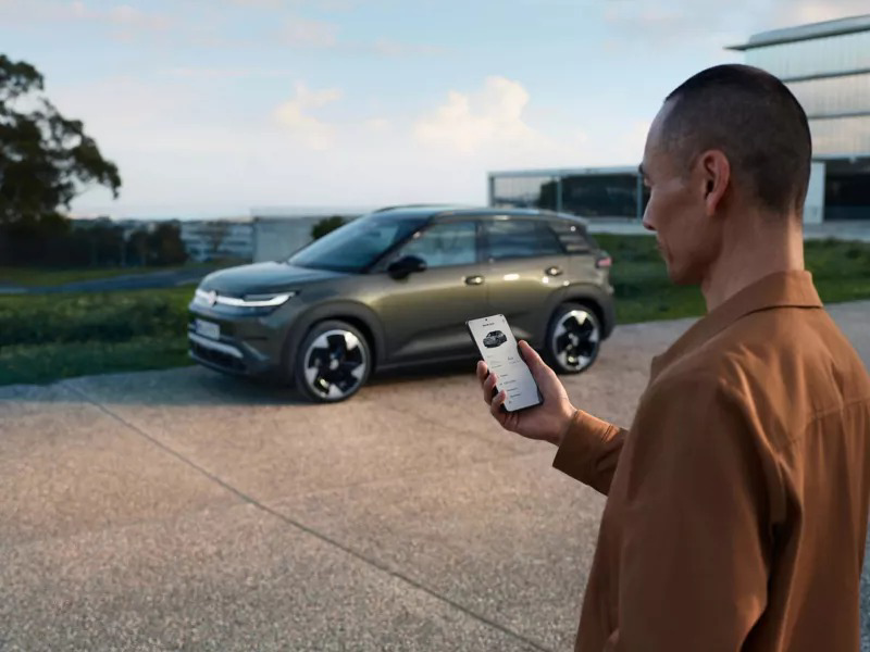 A man holding a smartphone with a car app, with a grey Volkswagen electric SUV parked in the background.