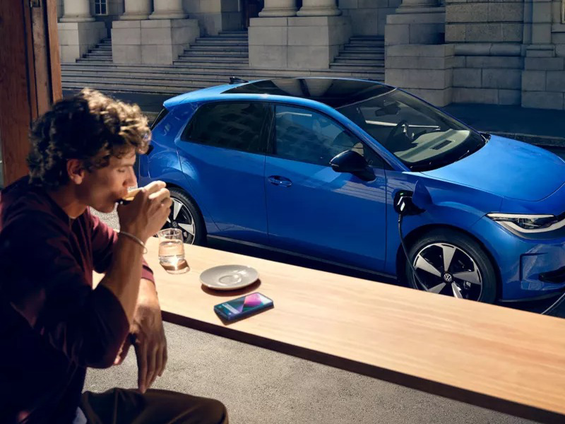 A man drinking coffee at an outdoor table while a blue Volkswagen electric hatchback charges in the background.
