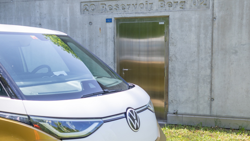 Un VW ID. Buzz se tient au premier plan. Derrière lui se trouve un bâtiment en béton avec une porte métallique.