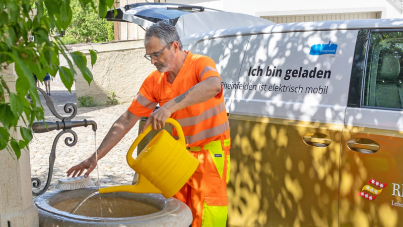Une personne en tenue de travail orange remplit un arrosoir jaune à une fontaine historique. À côté se trouve un VW ID. Buzz blanc et or portant l’inscription « Ich bin geladen – Rheinfelden ist elektrisch mobil ».