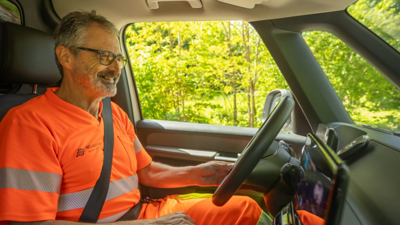 Eine Person in orangefarbener Arbeitskleidung sitzt lächelnd am Steuer eines Fahrzeugs. Durch das Seitenfenster sind grüne Bäume und Sonnenlicht zu sehen, der Innenraum wirkt modern und hell.