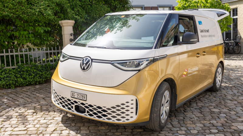 Un VW ID. Buzz bianco e oro con la dicitura di Rheinfelden è parcheggiato su una piazza lastricata. Una porta laterale e il portellone sono aperti, e gli alberi circostanti si riflettono sul parabrezza.