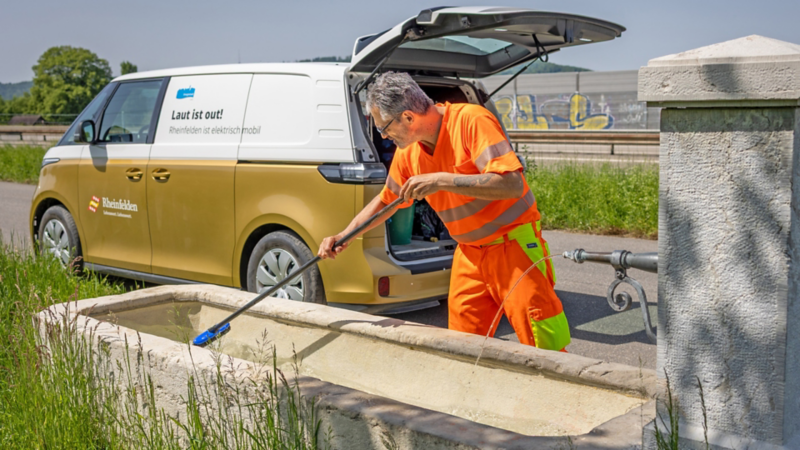 Eine Person in orangefarbener Arbeitskleidung reinigt mit einer langen Bürste ein Steintrog-Brunnenbecken. Im Hintergrund steht ein gold-weisser VW ID. Buzz der Stadt Rheinfelden mit offenem Kofferraum und der Aufschrift ‚Laut ist out! Rheinfelden ist elektrisch mobil‘.