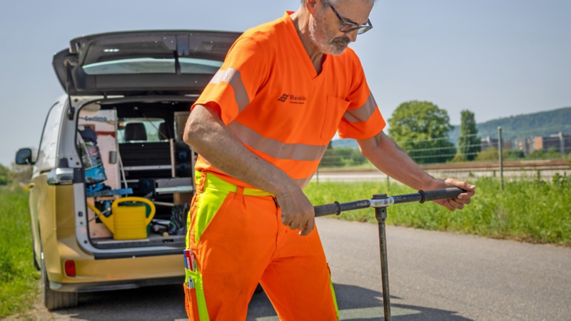 Eine Person in orangefarbener Arbeitskleidung bedient ein Werkzeug, während im Hintergrund der geöffnete Kofferraum eines gold-weissen Servicefahrzeugs mit diversen Arbeitsmaterialien sichtbar ist.