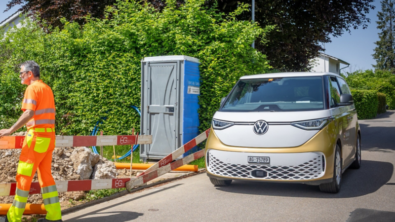 Une personne en tenue de travail orange passe devant un chantier fermé. À côté se trouve un VW ID. Buzz blanc et or portant la mention de Rheinfelden.