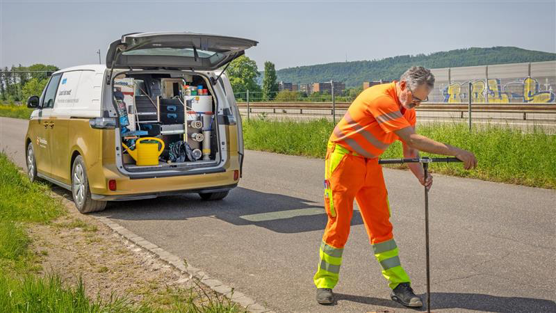 Eine Person in orangefarbener Arbeitskleidung bedient auf einer Strasse ein langes Werkzeug. Hinter ihr steht ein gold-weisser  VW ID. Buzz mit geöffnetem Kofferraum, in dem verschiedene Arbeitsmaterialien und Geräte sichtbar sind.