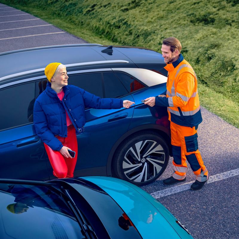 Skats no sāniem uz zilu VW ID.4, divi cilvēki sarunājas, priekšplānā redzama VW ID.3 aizmugure