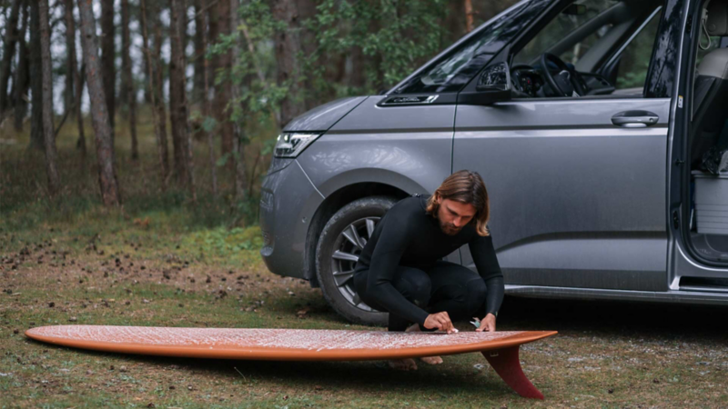 Sebastian slipar på sin surfbräda framför VW California husbil.