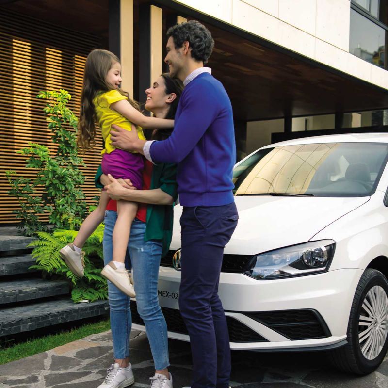 Familia de papá, mamá e hija afuera de una casa y delante de un auto sedán Vento Startline 2022. 