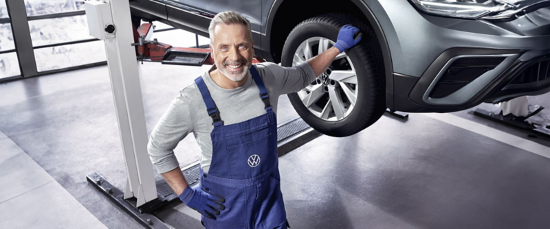 Un mécanicien Volkswagen souriant en salopette et gants bleus est debout à côté d'une voiture sur un pont élévateur dans un garage.