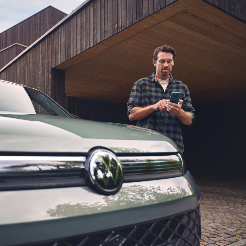 Close-up of a green Volkswagen car with a man in a plaid shirt looking at his phone in the background