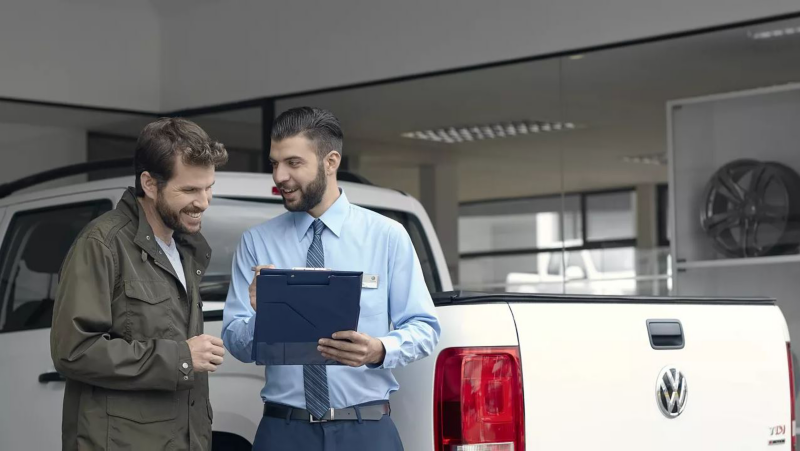 Un asesor de servicio o ventas, vestido con camisa y corbata, sostiene un portapapeles mientras conversa amablemente con un cliente. Detrás de ellos se ve la parte trasera de una pick-up Volkswagen Amarok blanca, sugiriendo una consulta o entrega de vehículo en un concesionario o taller.