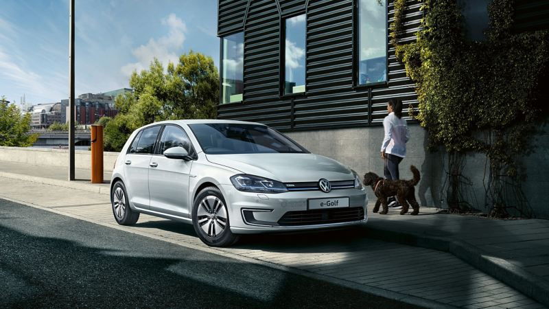 A woman walking a dog on a pavement alongside a parked e-Golf