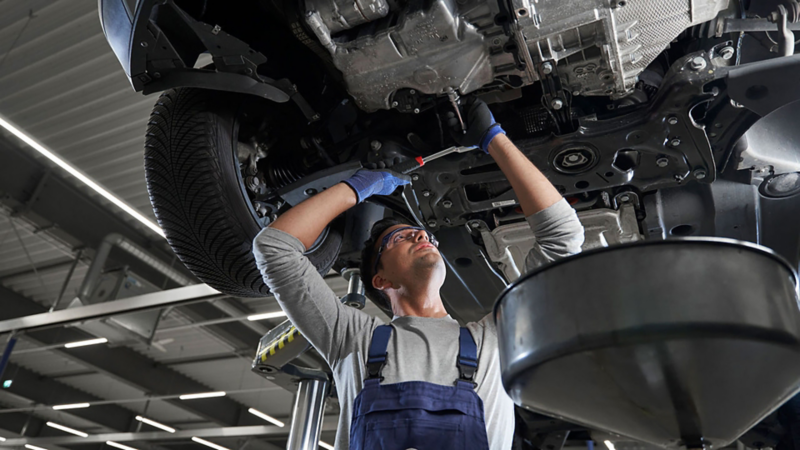 A Volkswagen mechanic underneath a car doing an oil inspection with the car up on the ramp
