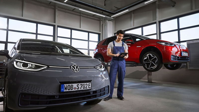 A Volkswagen mechanic between two Volkswagen cars in a Volkswagen service centre with one of the cars raised up on a ramp